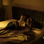 A restless man lying awake on his bed in a dimly lit bedroom at night.
