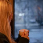 A woman with blonde hair relaxes indoors with a cup of coffee, looking out at the winter scene.