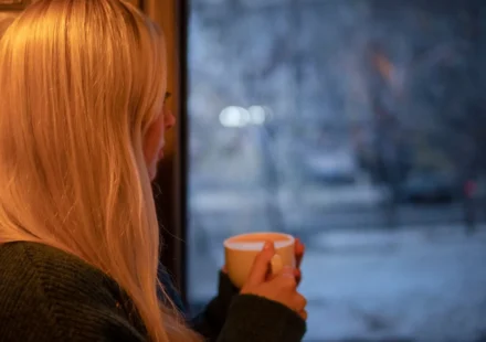 A woman with blonde hair relaxes indoors with a cup of coffee, looking out at the winter scene.