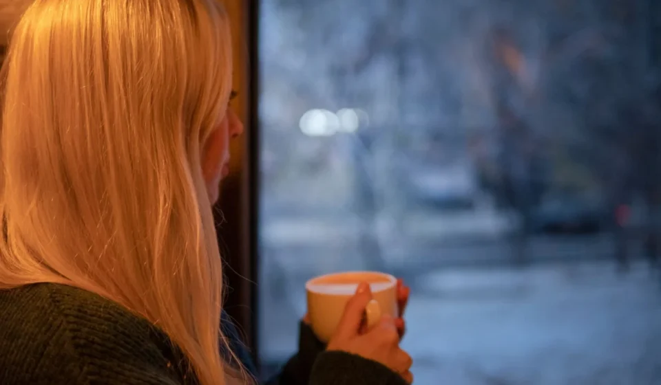 A woman with blonde hair relaxes indoors with a cup of coffee, looking out at the winter scene.