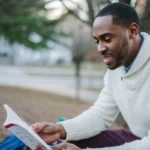 A young man enjoys reading in a serene park setting during the day.