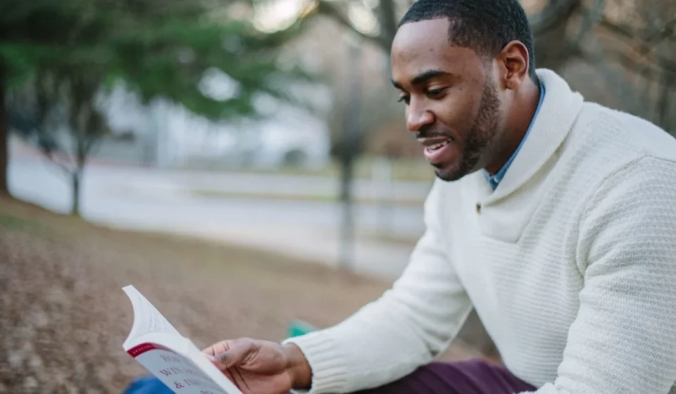 A young man enjoys reading in a serene park setting during the day.