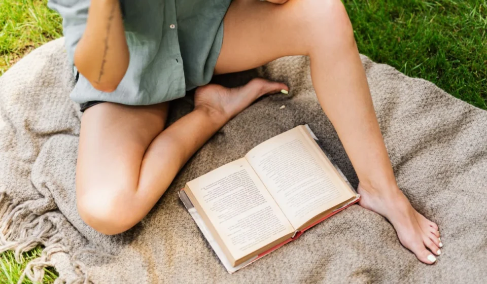 An adult enjoys a warm day outdoors, reading a book on a picnic blanket with bare feet.