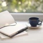 Cozy workspace setup with open notebook and coffee near a sunlit window.
