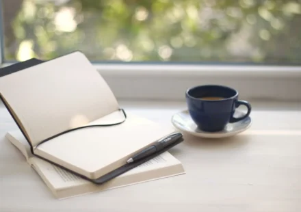 Cozy workspace setup with open notebook and coffee near a sunlit window.