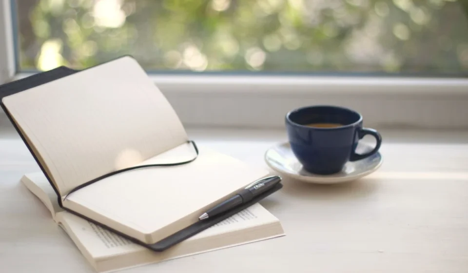 Cozy workspace setup with open notebook and coffee near a sunlit window.