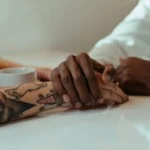 Interracial couple holding hands over coffee, showcasing love and connection with tattoos visible.