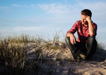 Man in a red plaid shirt sitting thoughtfully on a sandy dune in Australia, surrounded by grass.