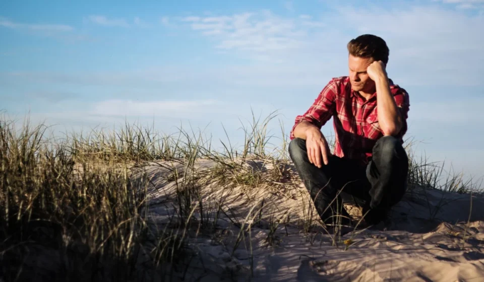 Man in a red plaid shirt sitting thoughtfully on a sandy dune in Australia, surrounded by grass.