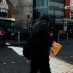 Man in city street scene holding a newspaper with urban buildings in background.