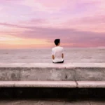 Man sitting alone by the ocean at sunset, embracing peace and tranquility.