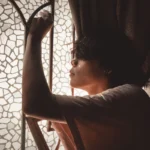 Side view of a young man posing thoughtfully by a mosaic glass window with soft light.