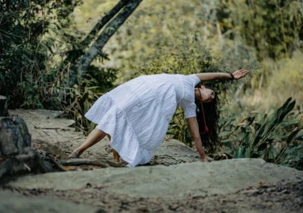 Woman in white dress practicing yoga on stone path in a lush forest setting.